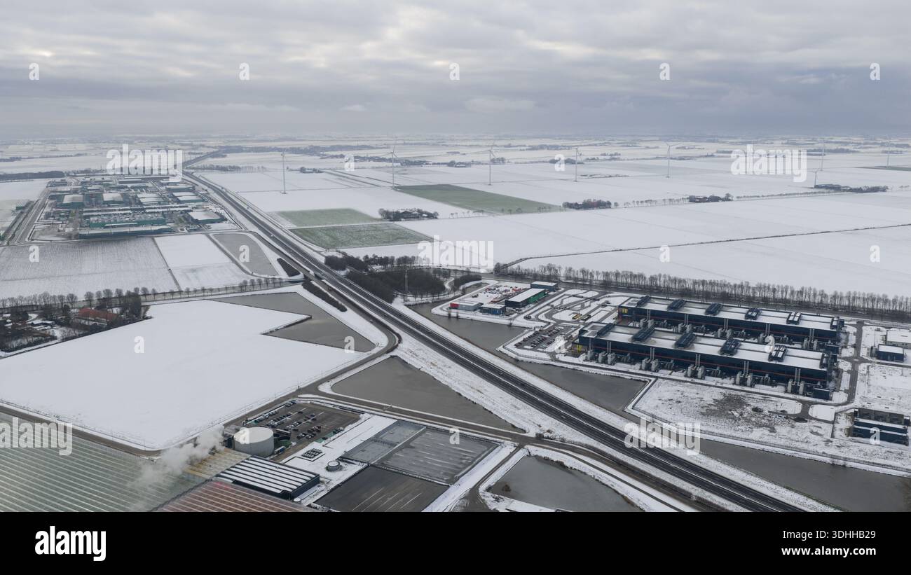 Aus der Vogelperspektive eine einfarbige Winterlandschaft, in der Schnee die flachen Felder und Gebäude bedeckt, im Kontrast zu den dunklen Straßen und Industriestrukturen Stockfoto