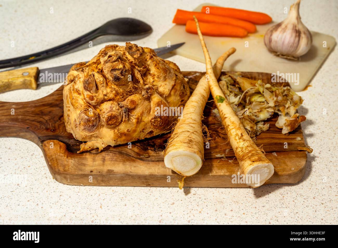 Halbierte Selleriewurzel und Petersilie auf Schneidebrett aus Olivenholz, Karotten und Zwiebeln, Löffel und Messer auf Küchentisch. Zubereitung der Mahlzeit. Stockfoto