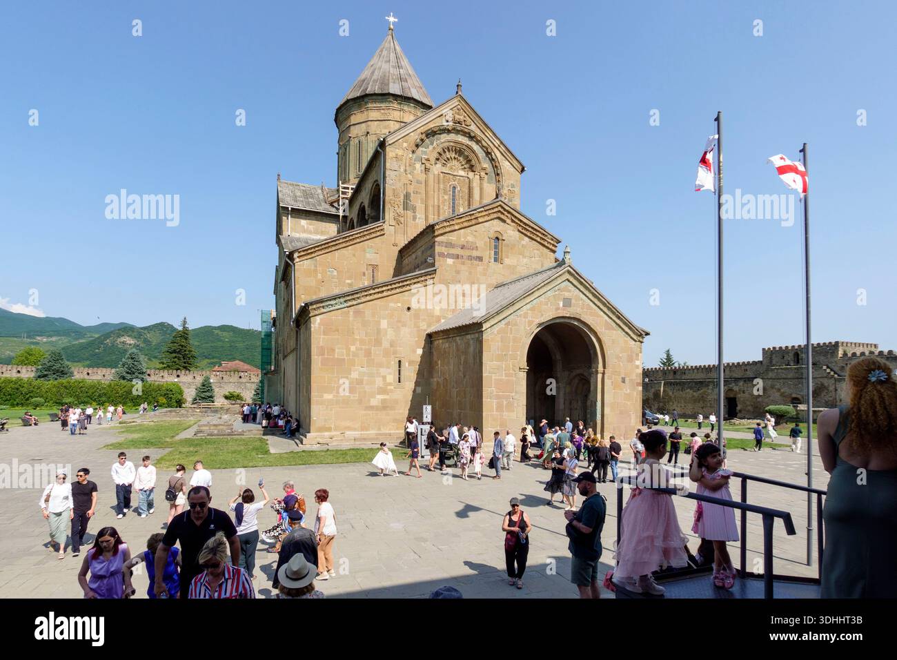 Vor der Svetitskhoveli-Kathedrale versammeln sich zahlreiche Besucher unter klarem Himmel mit georgianischen Fahnen, die im Vordergrund in Mzcheta, Georgien, schwenken Stockfoto