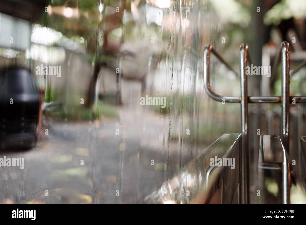 Eine geringe Tiefe der Feldaufnahme mit Fokus auf Wassertröpfchen, die über eine klare Glasoberfläche laufen, mit einem Bokeh-Effekt, der einen unscharfen Garten im Freien zeigt Stockfoto