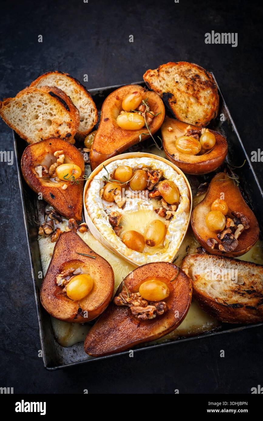 Im Ofen gebackener Camembert-Käse mit karamelisierten glasierten Birnen, Walnüssen und Trauben sowie Baguette auf rustikalem Metallteller Stockfoto