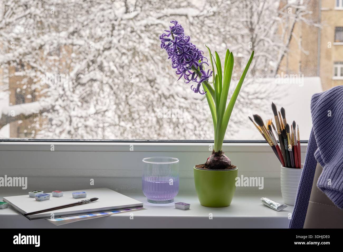 Eine gemütliche Fensterbank mit schneebedecktem Blick auf die Stadt, Kunstpinsel und Farben liegen bereit, damit ein Künstler eine Hyazinthe und eine urbane Landschaft malen kann. Stockfoto