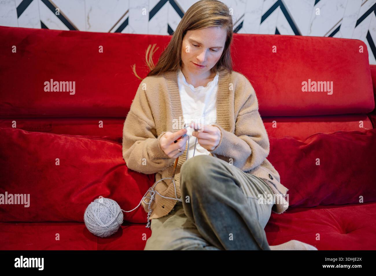 Eine Frau strickt, während sie auf einer roten Couch sitzt. Sie konzentriert sich auf ihr Handwerk mit einem Garnknäuel neben ihr in einem hellen Innenbereich. Stockfoto