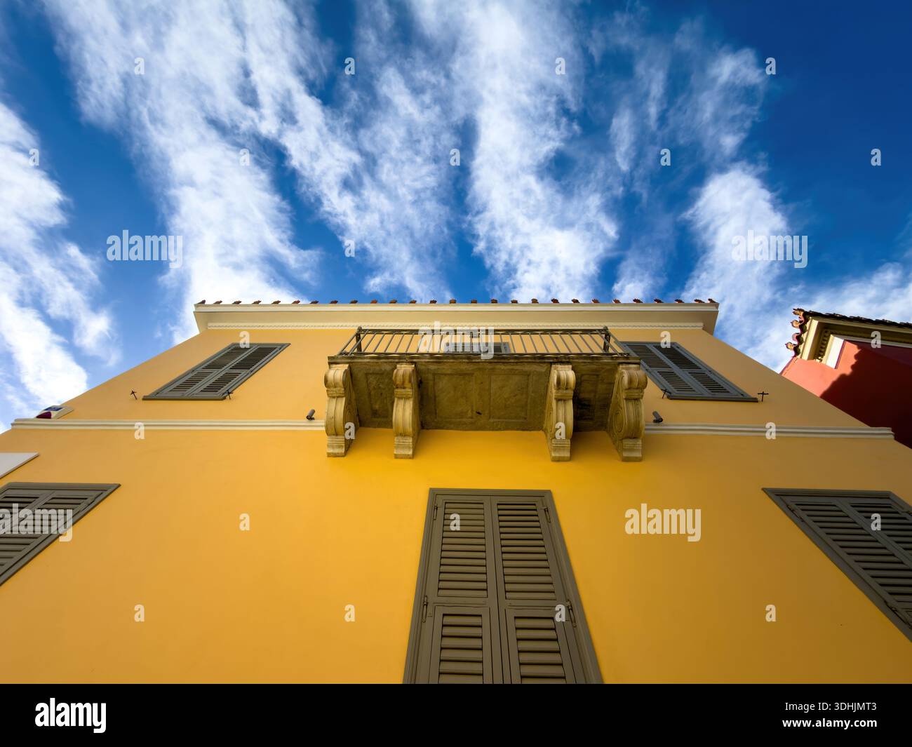 Gelbe neoklassizistische Gebäudefassade mit flachem Blick, Holzladenfenster und Marmorbalkon, blauer bewölkter Himmel. Plaka, Athen, Griechenland Stockfoto