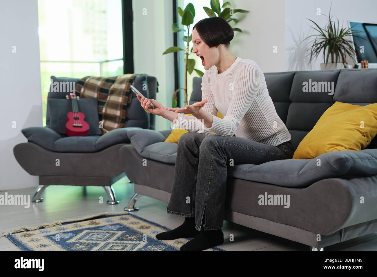 Frustration, die sichtbar ist, wenn eine junge Frau ihre Emotionen ausdrückt, während sie in einem gemütlichen Wohnzimmer ein Smartphone benutzt Stockfoto