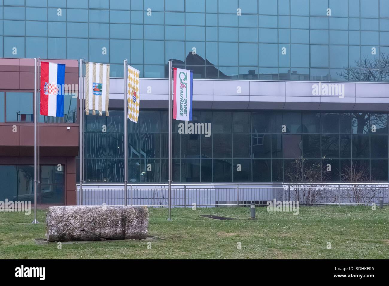 VINKOVCI, KROATIEN, 12.13.2025: Fahnen flattern an hohen Pfählen vor einem modernen Bürogebäude aus Glas und verleihen einem städtischen Außenbereich Farbe Stockfoto