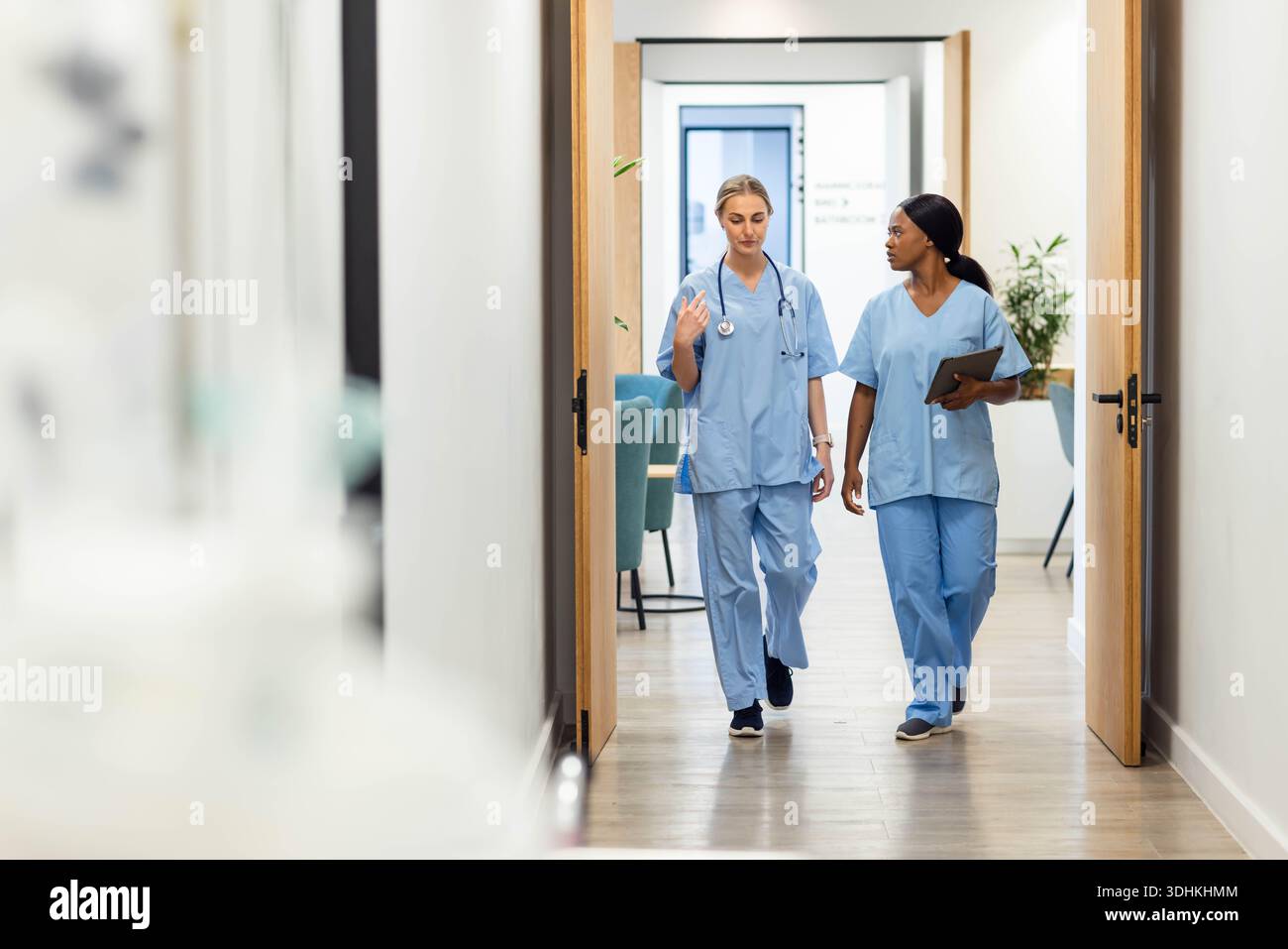 Diverse Krankenschwestern, die in blauen Peelings mit Stethoskop und Tablette den Korridor hinunterlaufen und sprechen Stockfoto