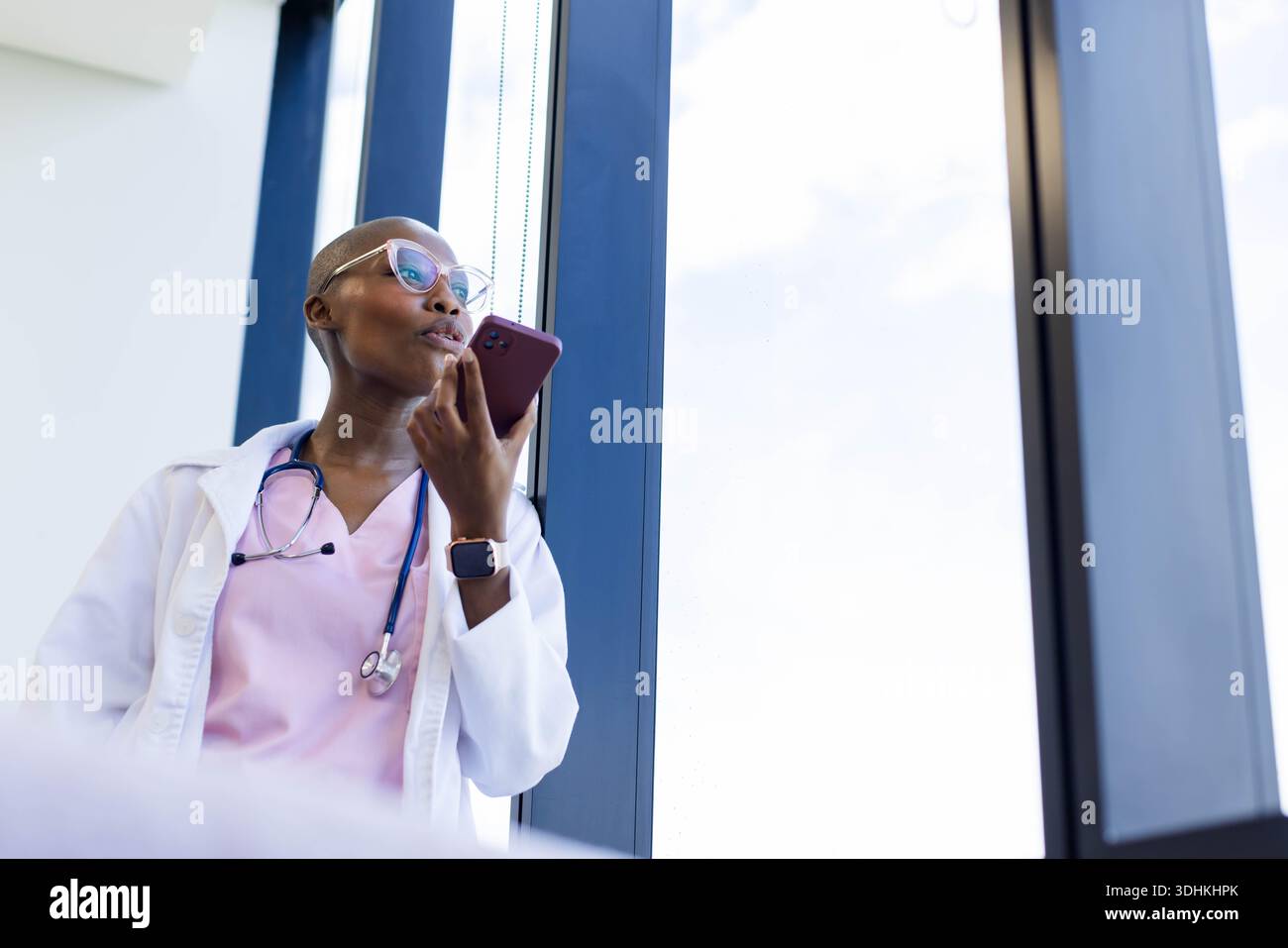 Afrikanische Ärztin, die am Klinikfenster steht, Telefon im Mantel und Stethoskop hält, Kopierraum Stockfoto