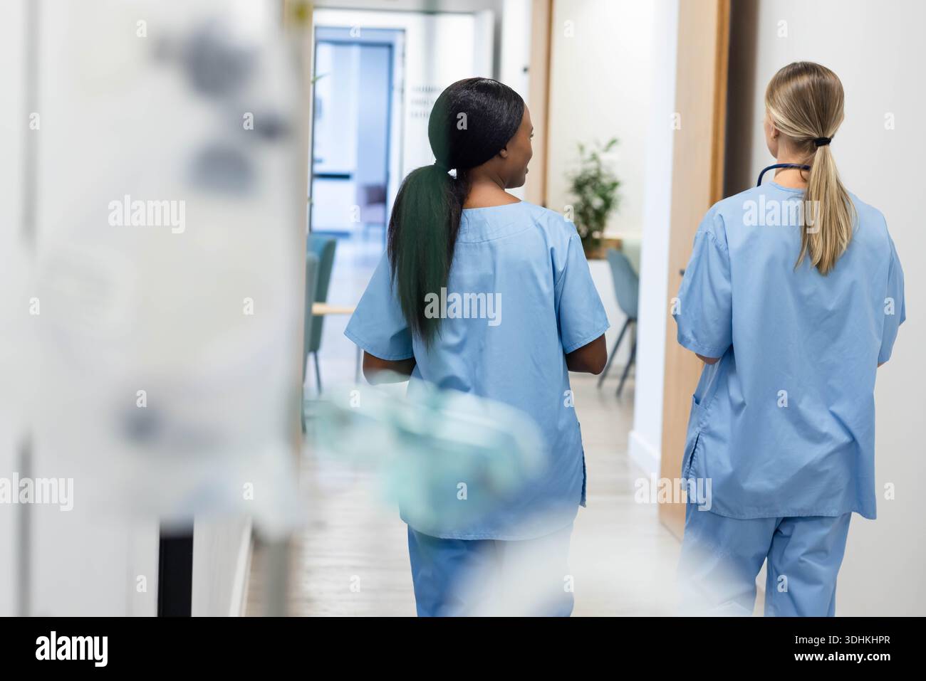 Zwei Ärztinnen, die den hellen Klinikkorridor in blauen Schutten hinunterlaufen, eine mit Stethoskop Stockfoto