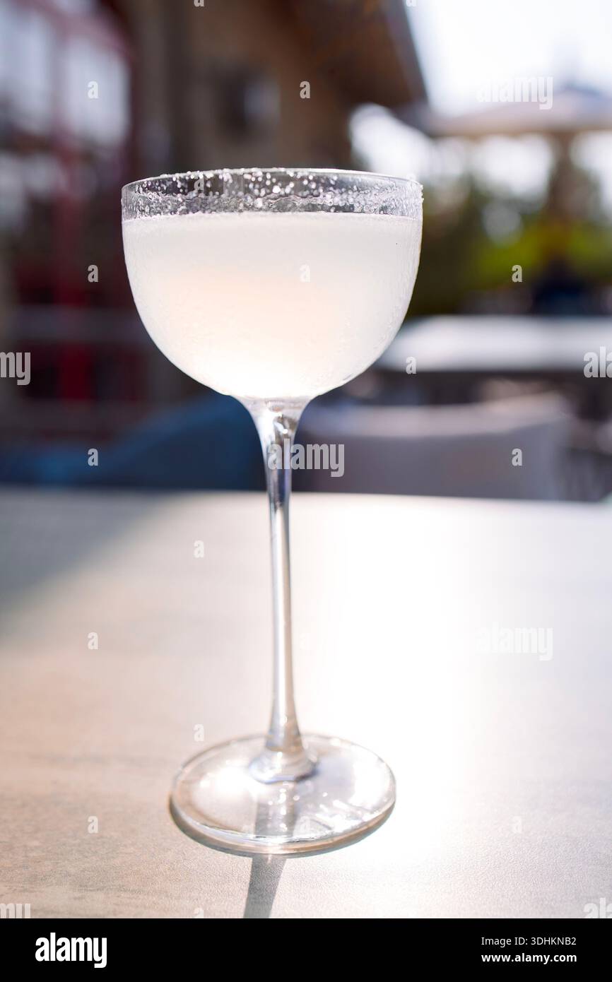 Das Cocktailglas bietet ein erfrischendes weißes Margarita-Getränk mit gesalzenem Rand, das auf einem Tisch in lebhafter Atmosphäre im Freien steht und ideal für gesellschaftliche Veranstaltungen ist Stockfoto