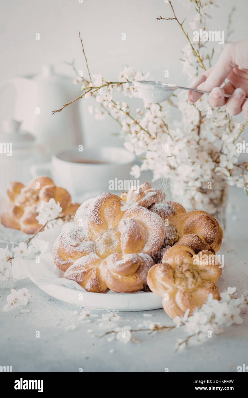 Ein Nahbild von wunderschön gebackenem Gebäck, das mit Puderzucker bestäubt ist, begleitet von einer Vase mit weißen Blumen und einer Hand, die die Blumen anpasst. Th Stockfoto