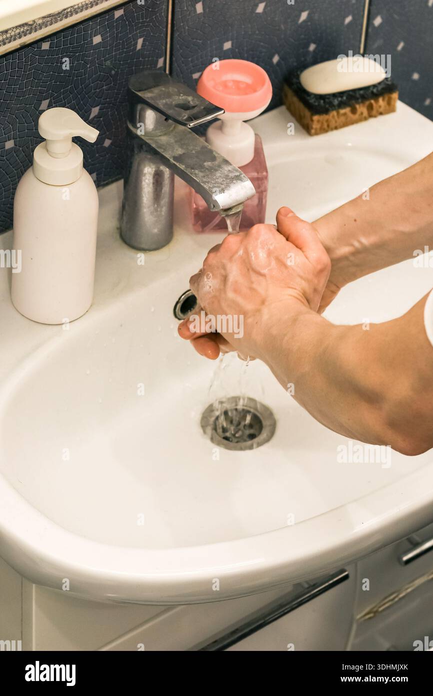 Einzelne seifige Hände unter dem Wasserhahn in einem Waschbecken mit gekacheltem Hintergrund aneinander reiben Stockfoto