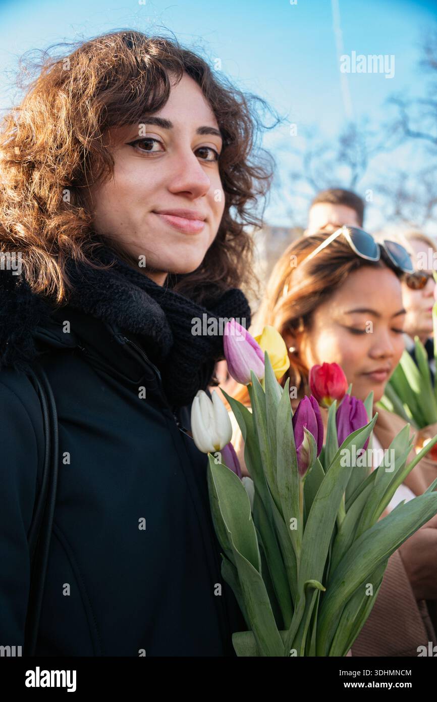 Eine lächelnde Frau mit lockigen Haaren, die in die Kamera schaut, hält einen mehrfarbigen Tulpenstrauß am Amsterdamer Tulpentag, an einem sonnigen Wintertag Stockfoto