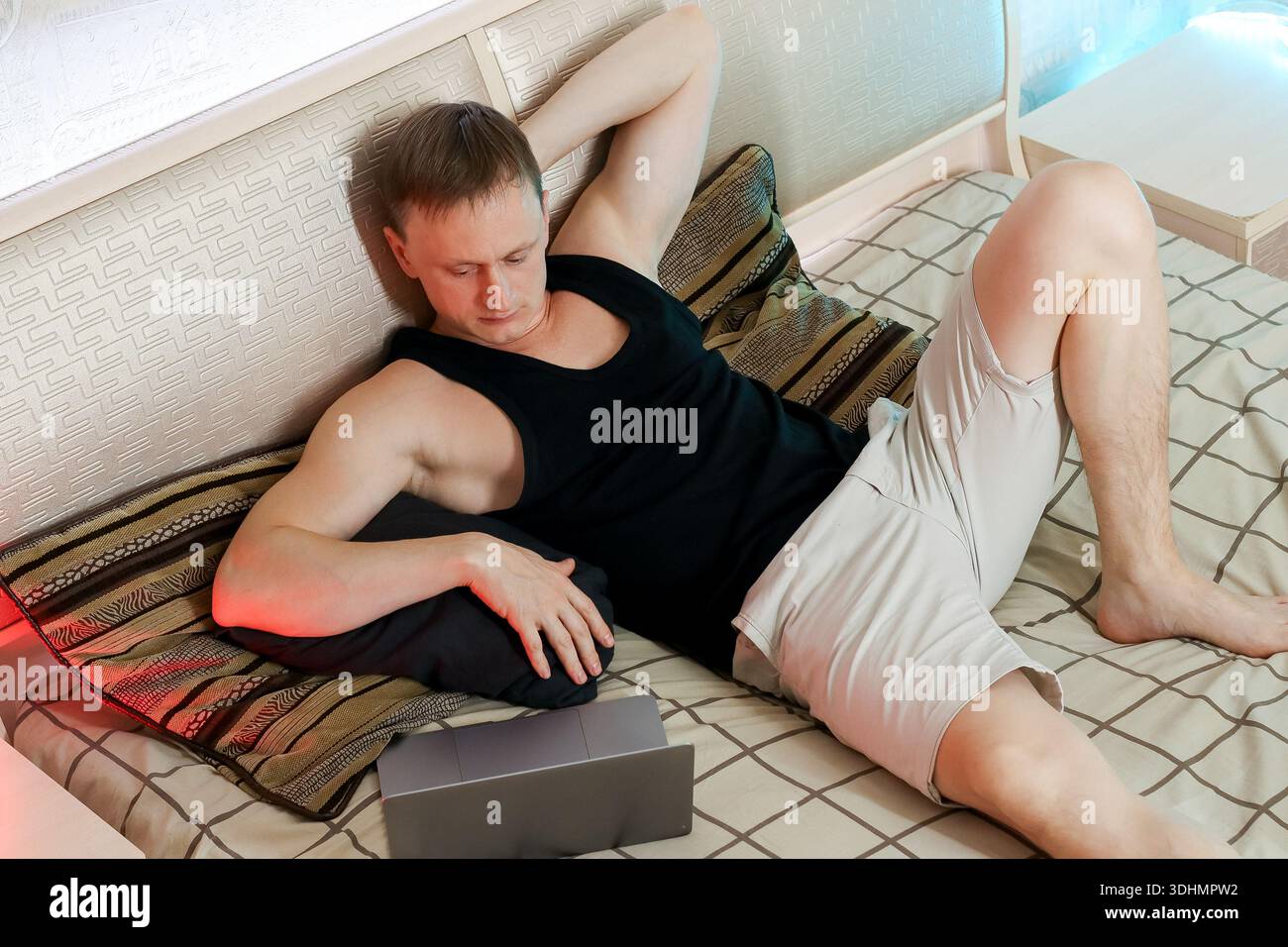 Auf einem gemusterten Bett mit ausgestrecktem Arm liegt ein Mann in lässiger Kleidung neben seinem Laptop, umgeben von der warmen, diffusen Beleuchtung einer Person Stockfoto