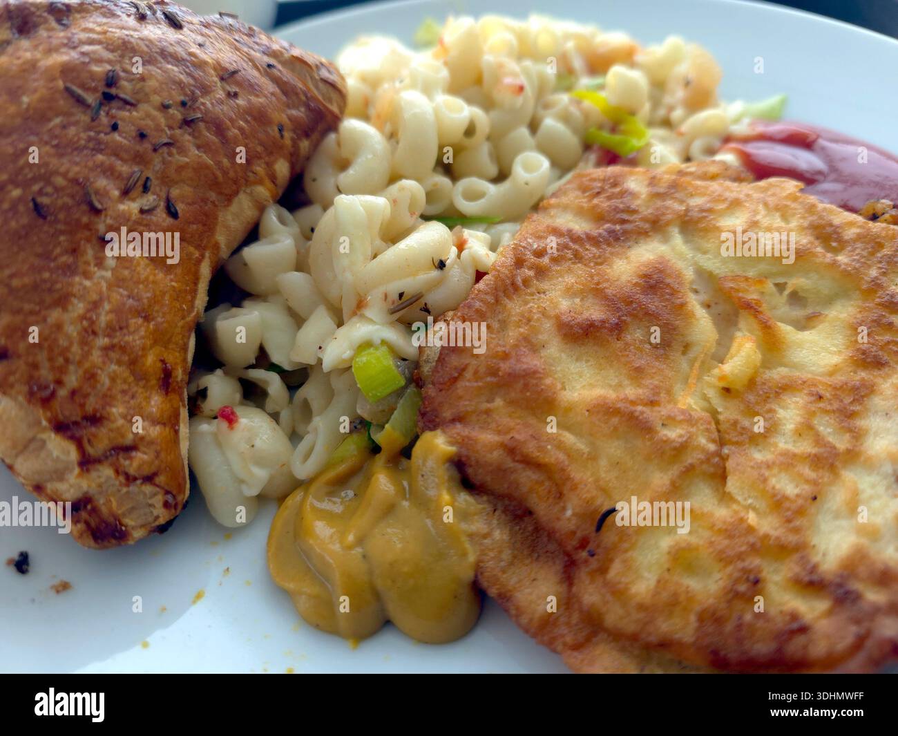 Ein Teller bietet eine Mischung aus Speisen wie Pasta, Gebäck und ein frittiertes Gericht zusammen mit Senf und Ketchup. Diese Mahlzeit wird mittags genossen. Stockfoto