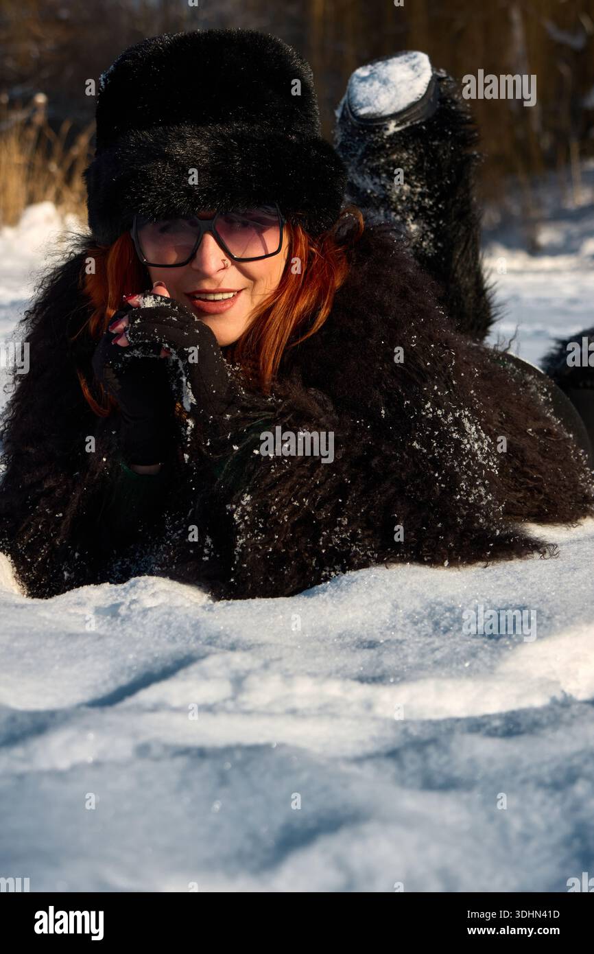 Eine schöne rothaarige Frau liegt im Winter auf dem Schnee, trägt einen luxuriösen Pelzmantel und einen passenden Pelzhut. Sie lächelt, ihre Augen funkeln vor Stockfoto