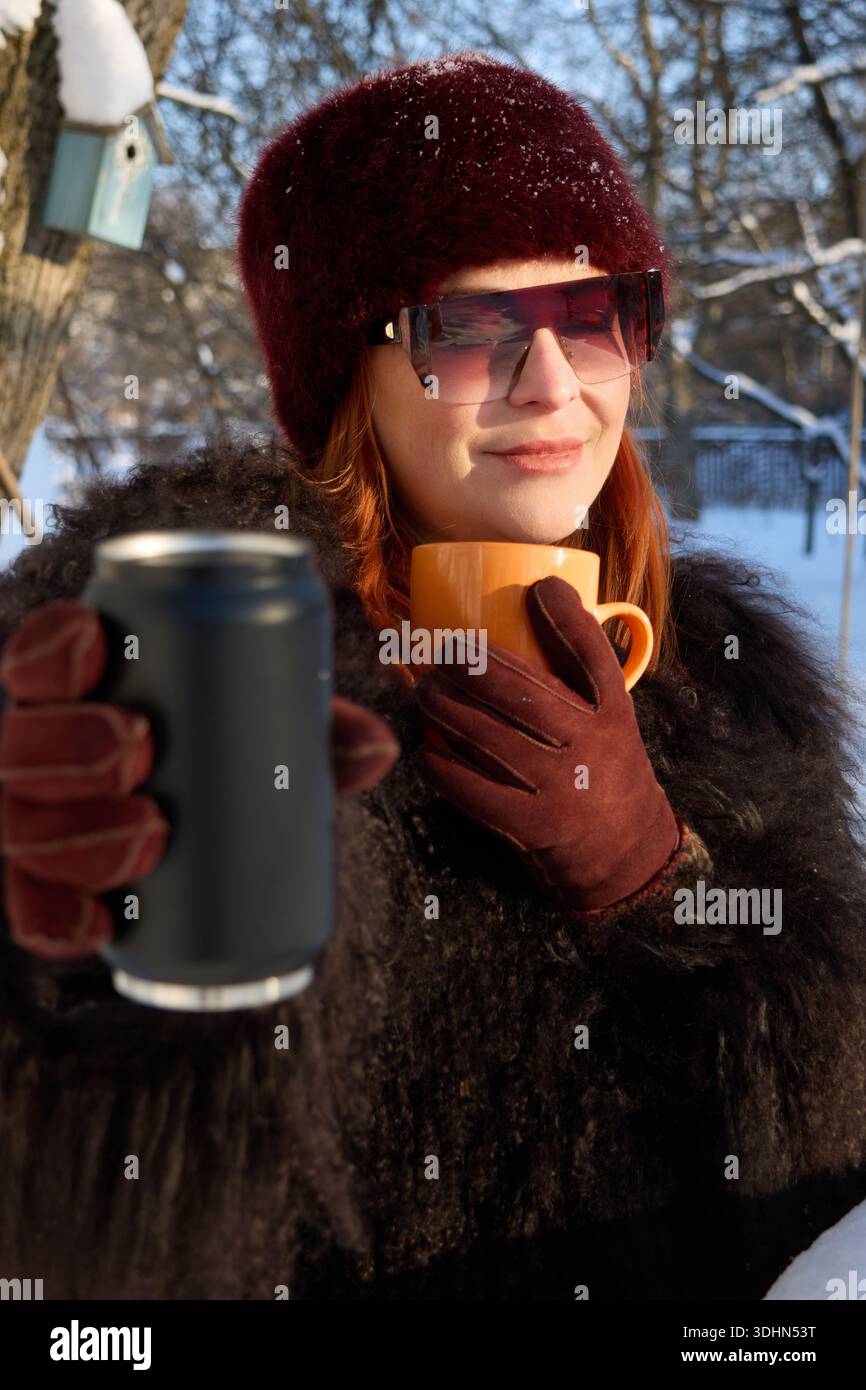 Dieses Bild zeigt eine stilvolle Frau mit leuchtend roten Haaren, gekleidet in einem dunklen, strukturierten Pelzmantel, einem burgunderroten Pelzhut, der mit Schneeflocken bestäubt ist, und einem modischen Fellmantel Stockfoto