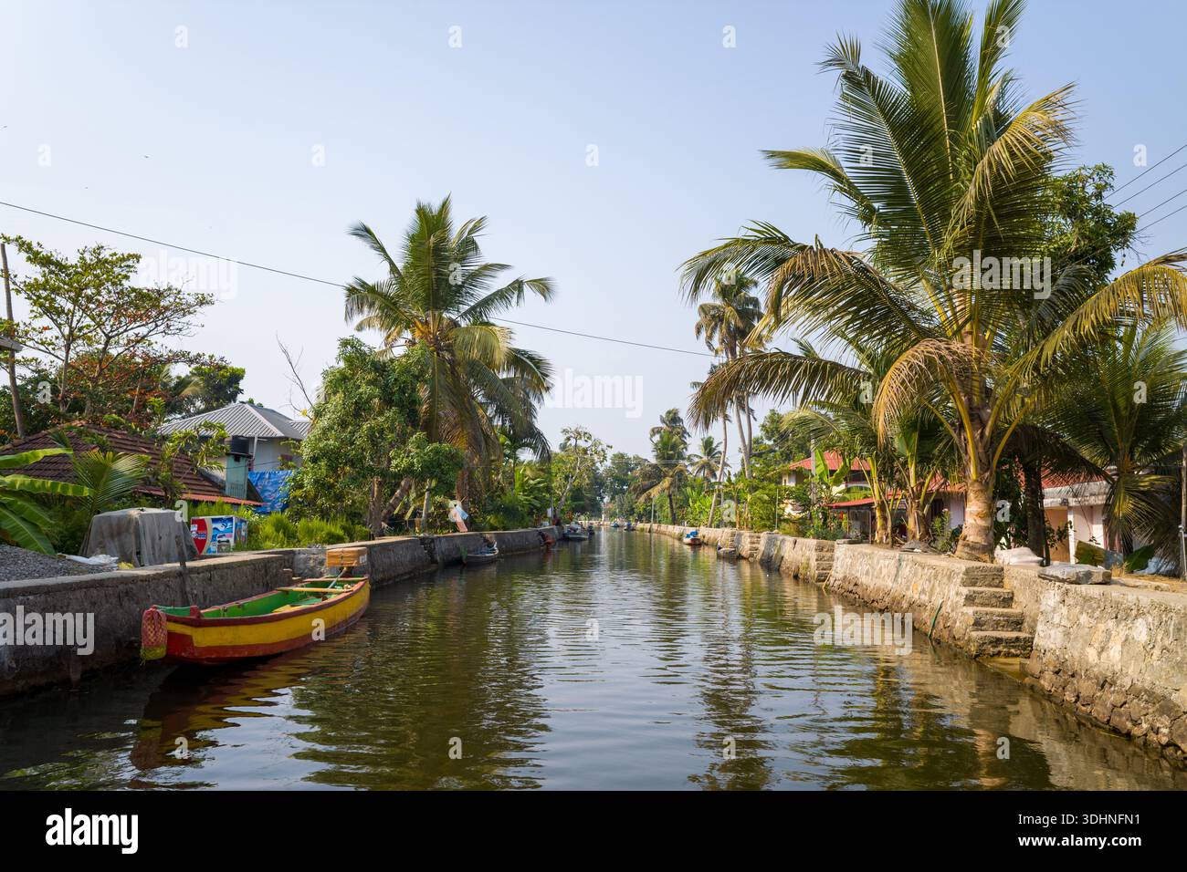 Alappuzha, Indien - 7. März 2023: Ein Kanal, gesäumt von Steinböcken, Palmen und Wohnhäusern, mit einem Holzboot, das am Wasserrand unter klarem Himmel vertäut ist. Stockfoto