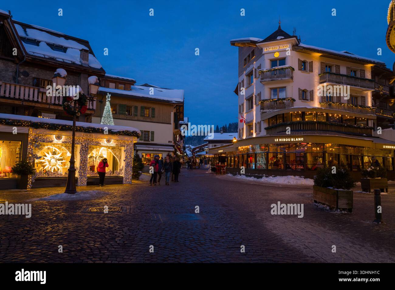 Megeve, Frankreich - 30. Januar 2023: Fußgänger spazieren am Abend in Megeve entlang einer kopfsteingepflasterten Straße mit beleuchteten Geschäften und dekorierten Gebäuden, darunter Le Hibou Blanc und Dior. Stockfoto