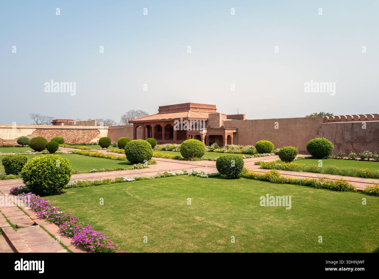 Fatehpur Sikri, Indien - 12. März 2024: Das Bild zeigt den roten Sandstein Diwan-E-AAM-Publikumssaal des Jodha Bai Palastkomplexes mit gepflegten Rasenflächen, Blumenbeeten und getrimmten Büschen im Vordergrund unter klarem Himmel. Stockfoto