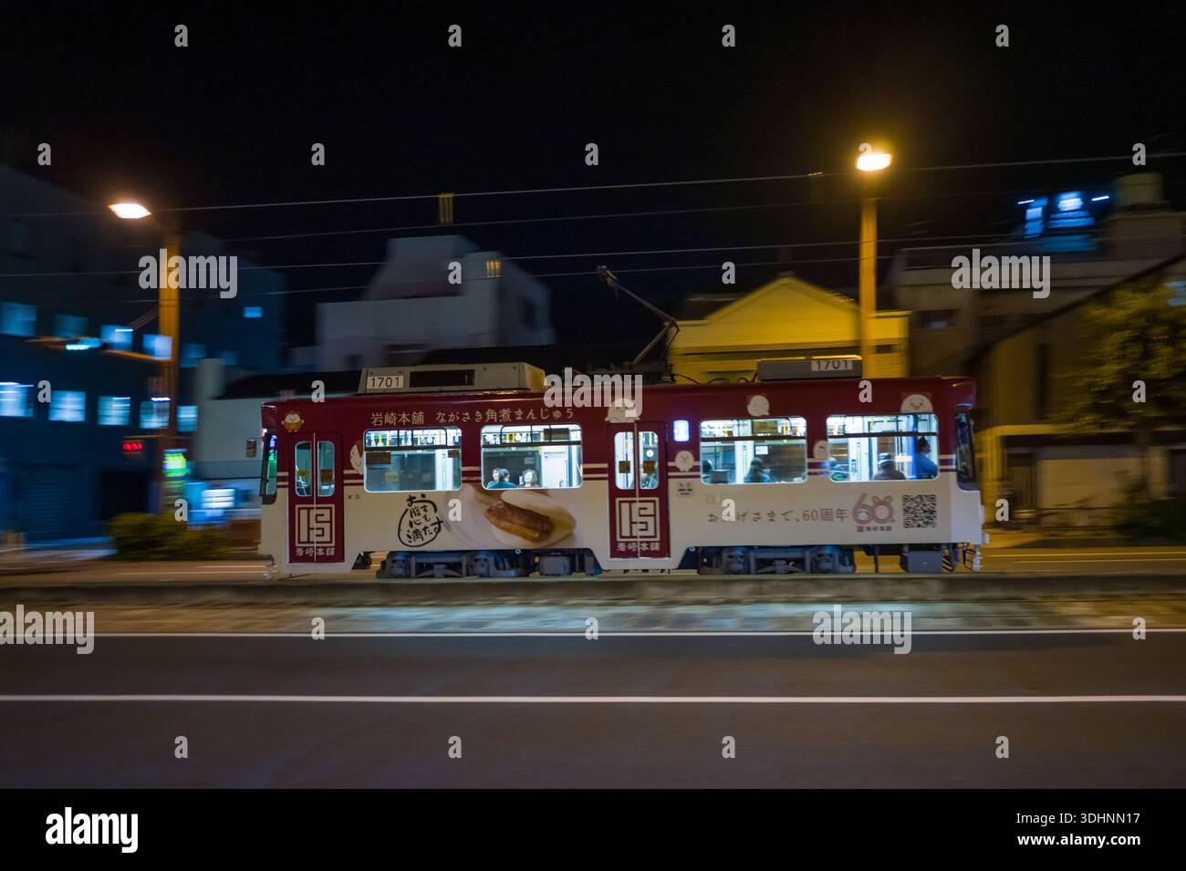 Nagasaki, Japan - 11. November 2024: Eine rot-weiße Straßenbahn mit der Nummer 1701 fährt nachts mit Passagieren, die durch beleuchtete Fenster sichtbar sind, mit japanischer Werbung für eine Süßwarenfabrik und einer Feier zum 60. Jahrestag. Stockfoto