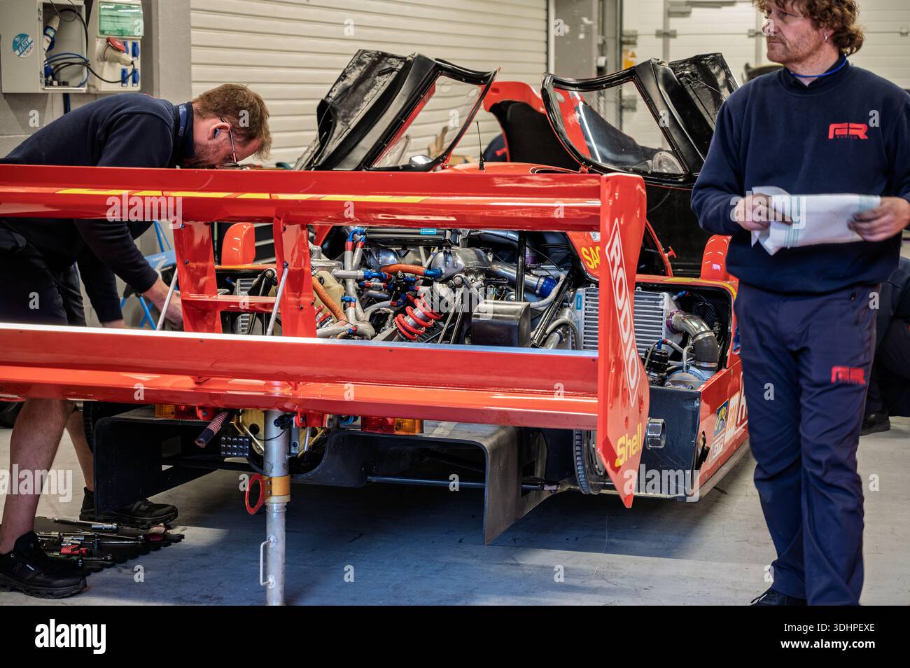 Rückansicht eines klassischen deutschen Porsche 962 C-Rennwagens aus dem Jahr 1992 in der Boxengarage, der von Mechanikern mit freiliegendem Motor vorbereitet wird Stockfoto