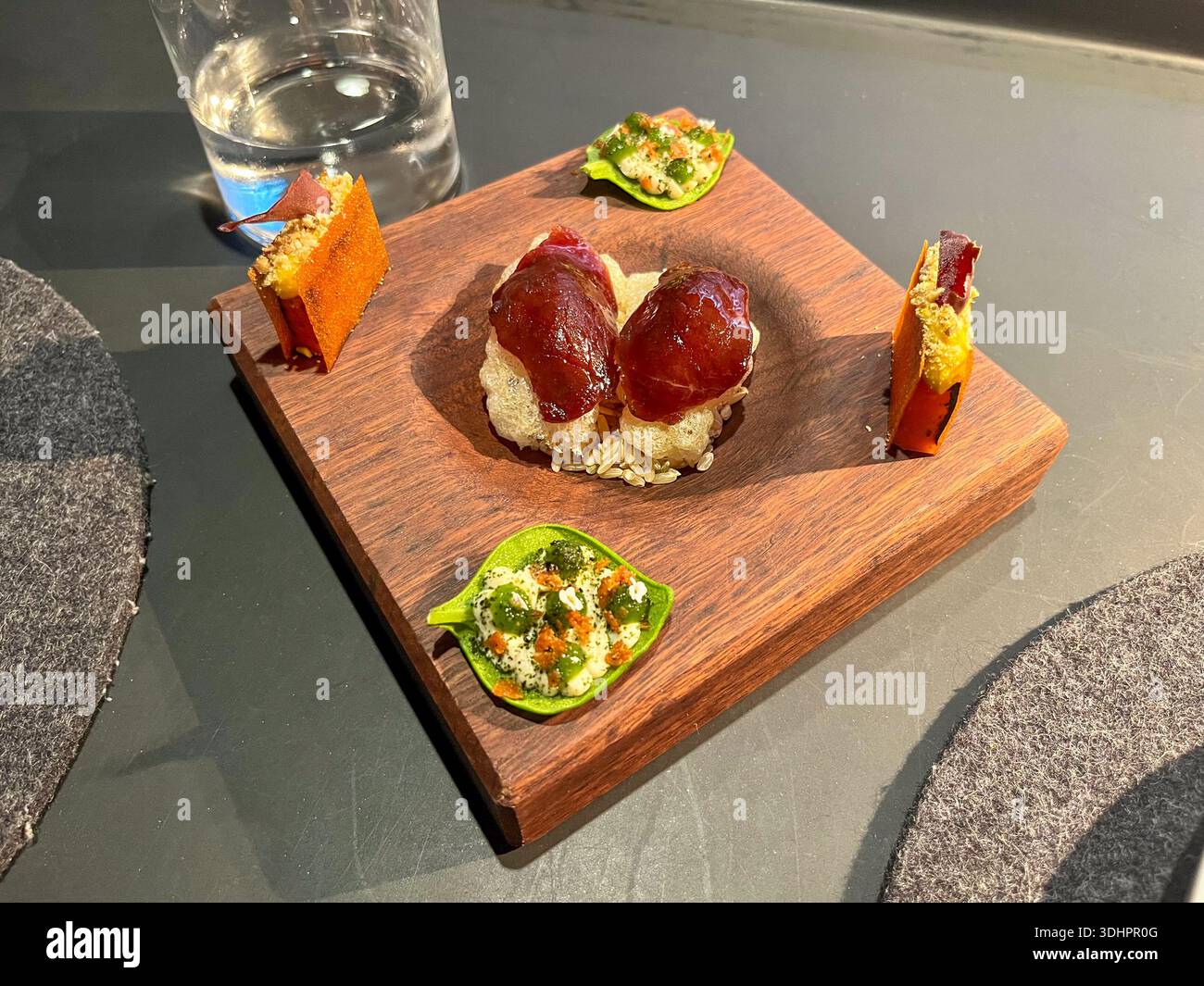 Fünf verschiedene herzhafte Vorspeisen und Amuse-Bouche sind kunstvoll auf einem dunklen Holzbrett angeordnet, zusammen mit einem erfrischenden Glas Wasser. Stockfoto