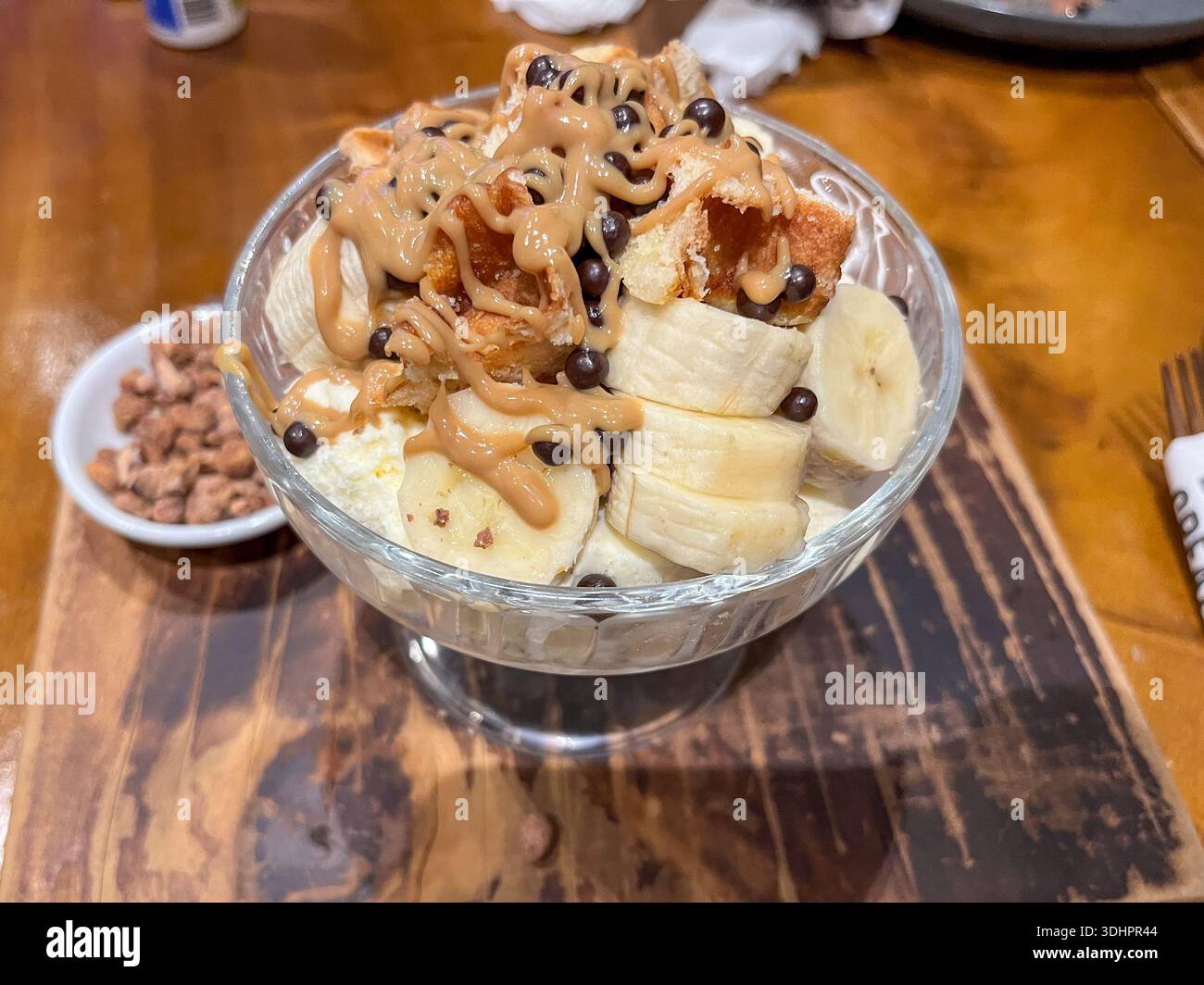 Ein cremiges Dessert in einer Glasschale bietet frische Bananenscheiben, knuspriges Brot, reichhaltige Erdnusssauce und kleine Schokoladenchips. Stockfoto
