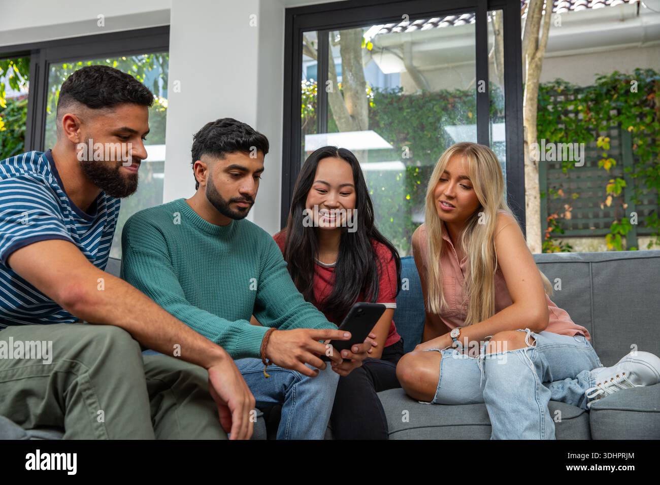 Verschiedene Freunde, die sich hineinlehnen und das Smartphone auf der grauen Couch im hellen Wohnzimmer ansehen Stockfoto