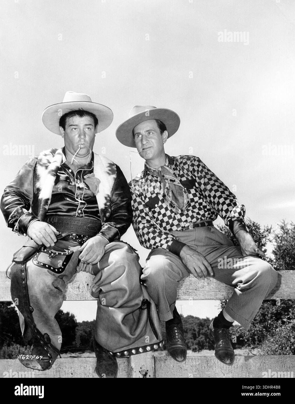 Aus den 1940er Jahren zeigt dieses Schwarz-weiß-Duo noch immer das legendäre Comedy-Duo Abbott und Costello, das in Cowboykleidung gekleidet ist und mit Strohhalmen im Mund auf einem Zaun sitzt. Lou Costello (links) trägt ein Satinhemd mit einer Pelzweste und Chaps, während Bud Abbott (rechts) ein kariertes Hemd und einen Schal trägt, sowohl nach oben als auch nach rechts, auf einem Filmset für ihren Film „Ride 'EM Cowboy“. Stockfoto