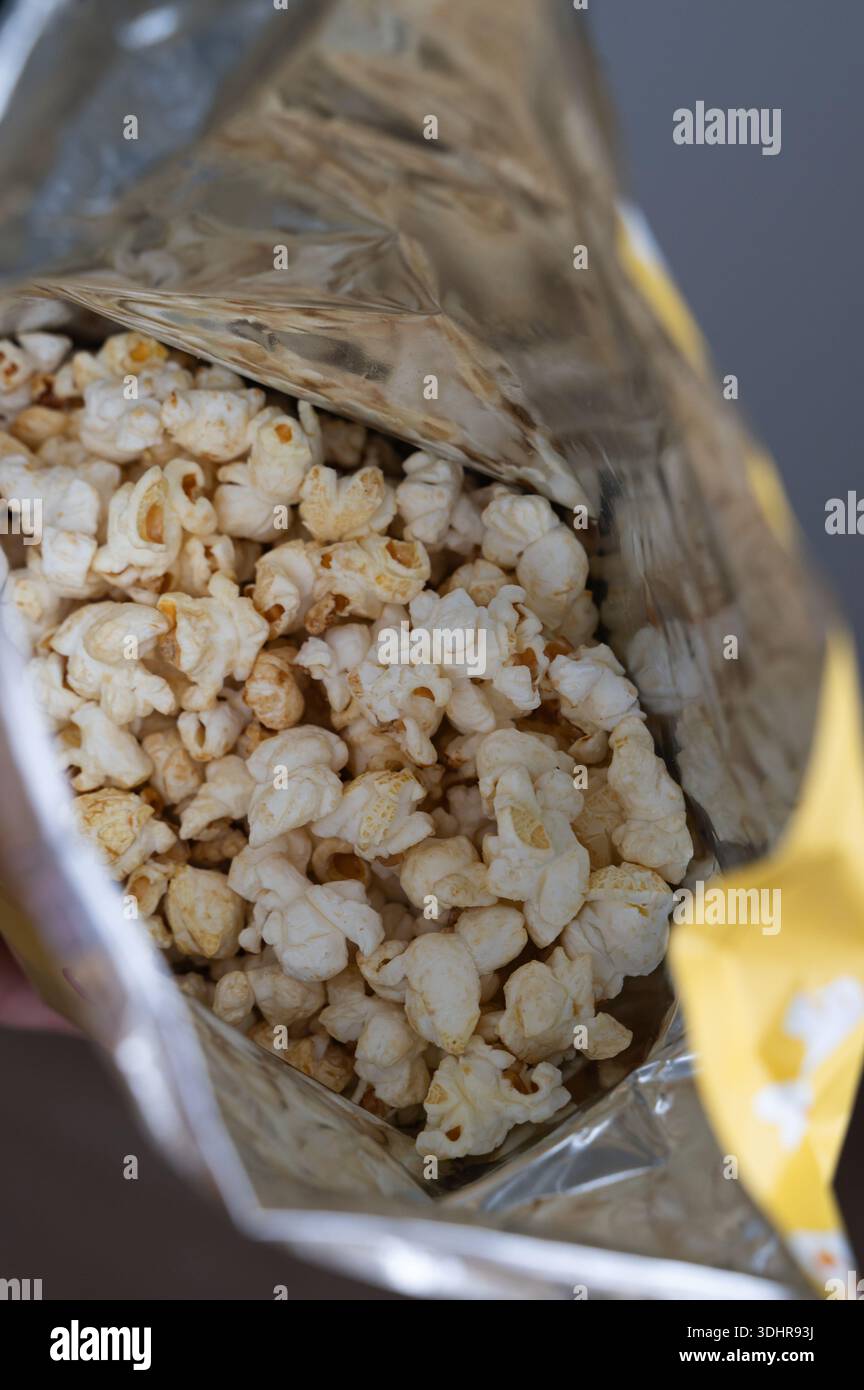 Offene Metalltüte Popcorn innen, Nahansicht mit gesalzenen Körnern und Folienverpackungen, gelber Brandmarkenrand, strukturierten Popcorn-Oberflächen, Inviti Stockfoto