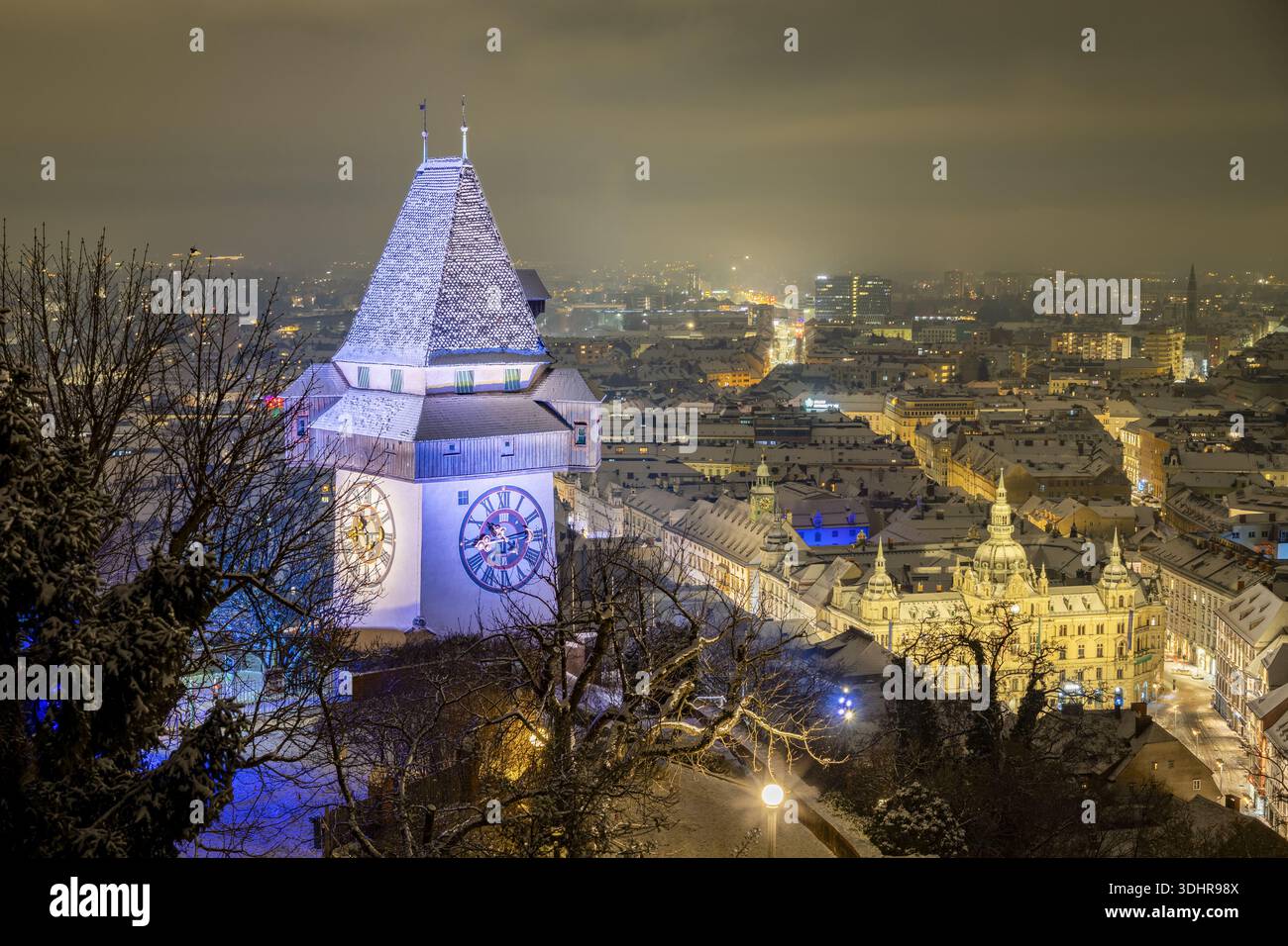 Blick auf den Uhrturm, der majestätisch gegen das Stadtbild steht, sein Gesicht beleuchtet, im Kontrast zum dunklen Himmel und schneebedeckten Bäumen, Graz, Stockfoto
