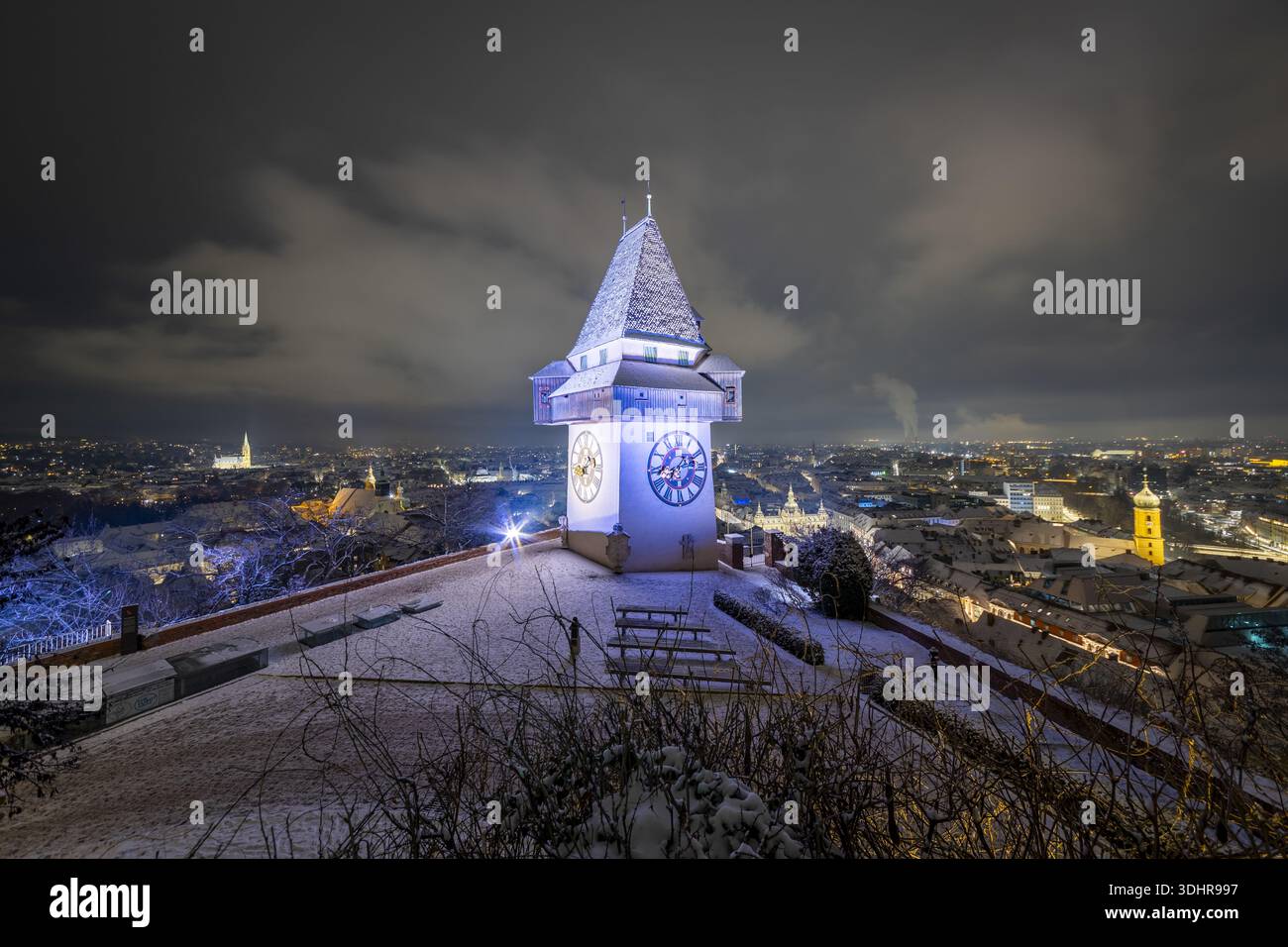 Der Blick auf den beleuchteten Glockenturm auf dem Schlossberg erhebt sich majestätisch gegen den Nachthimmel, mit dem verschneiten Stadtbild darunter, Graz, Stall Stockfoto