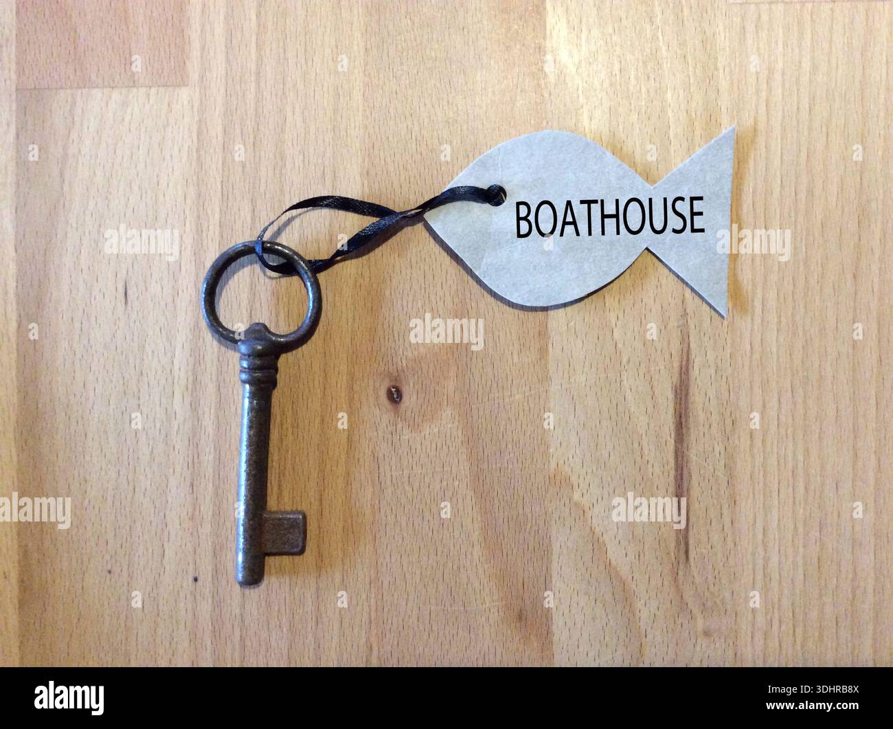 Antik verzierter Schlüssel, der an einem fischförmigen Tag mit der Aufschrift „BOAT HOUSE“ auf einer natürlichen Holzoberfläche hängt – ideales Symbol für ein Haus am Wasser Stockfoto