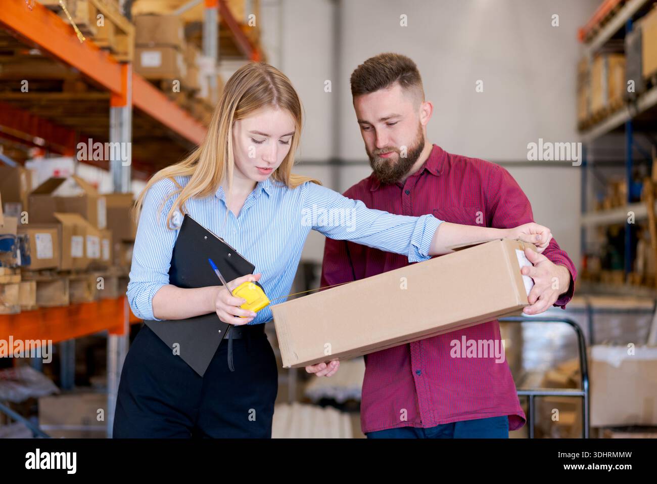 Emale Supervisor und männlicher Arbeiter messen einen Karton mit einem Maßband in einem Lager. Professionelles Team, das die Verpackungsabmessungen für die Untersuchung überprüft Stockfoto