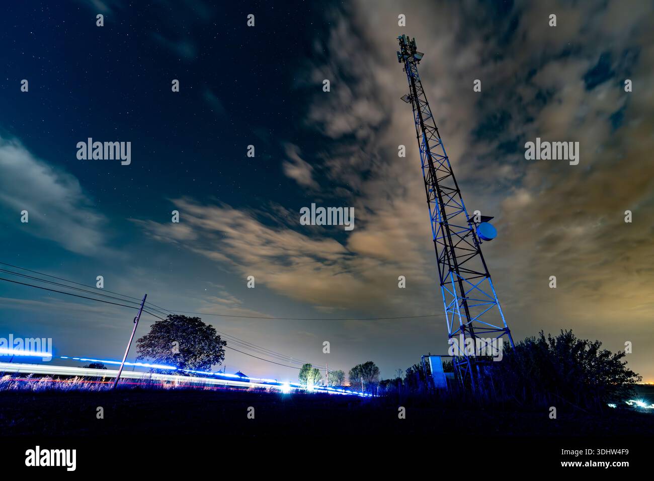 Ein Mobilfunkturm steht hoch am Nachthimmel. Wolken bewegen sich langsam, während ein Fahrzeug vorbeifährt, und hinterlassen leichte Spuren. Sterne sind im Hintergrund sichtbar Stockfoto