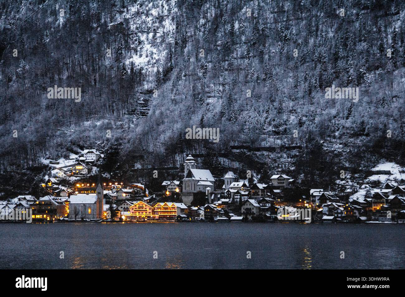 Gemütliches europäisches Bergdorf mit schneebedeckten Dächern und warmen Lichtern am Fuße eines satinierten Bergwaldes an einem ruhigen See in der Dämmerung. Stockfoto