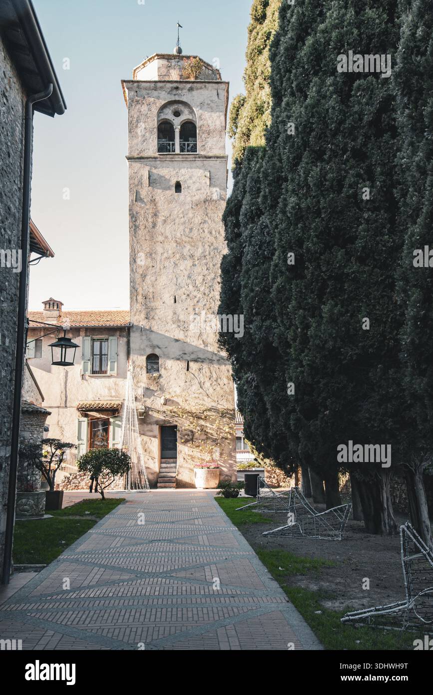 Blick auf einen Steinweg, der zu einem verwitterten Turm mit hohen, dunkelgrünen Bäumen führt, die einen natürlichen Rahmen in Sirmione, Lombardei, Italien bilden. Stockfoto