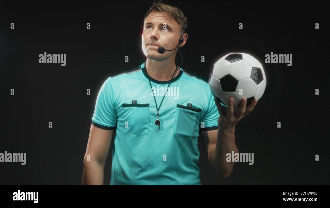 Der Schiedsrichter hält den Fußball in der rechten Hand, trägt eine Pfeife auf dem Schlüsselband und dem Headset-Mikrofon, während er im Studio nach oben blickt; durchdachte Autorität. Stockfoto