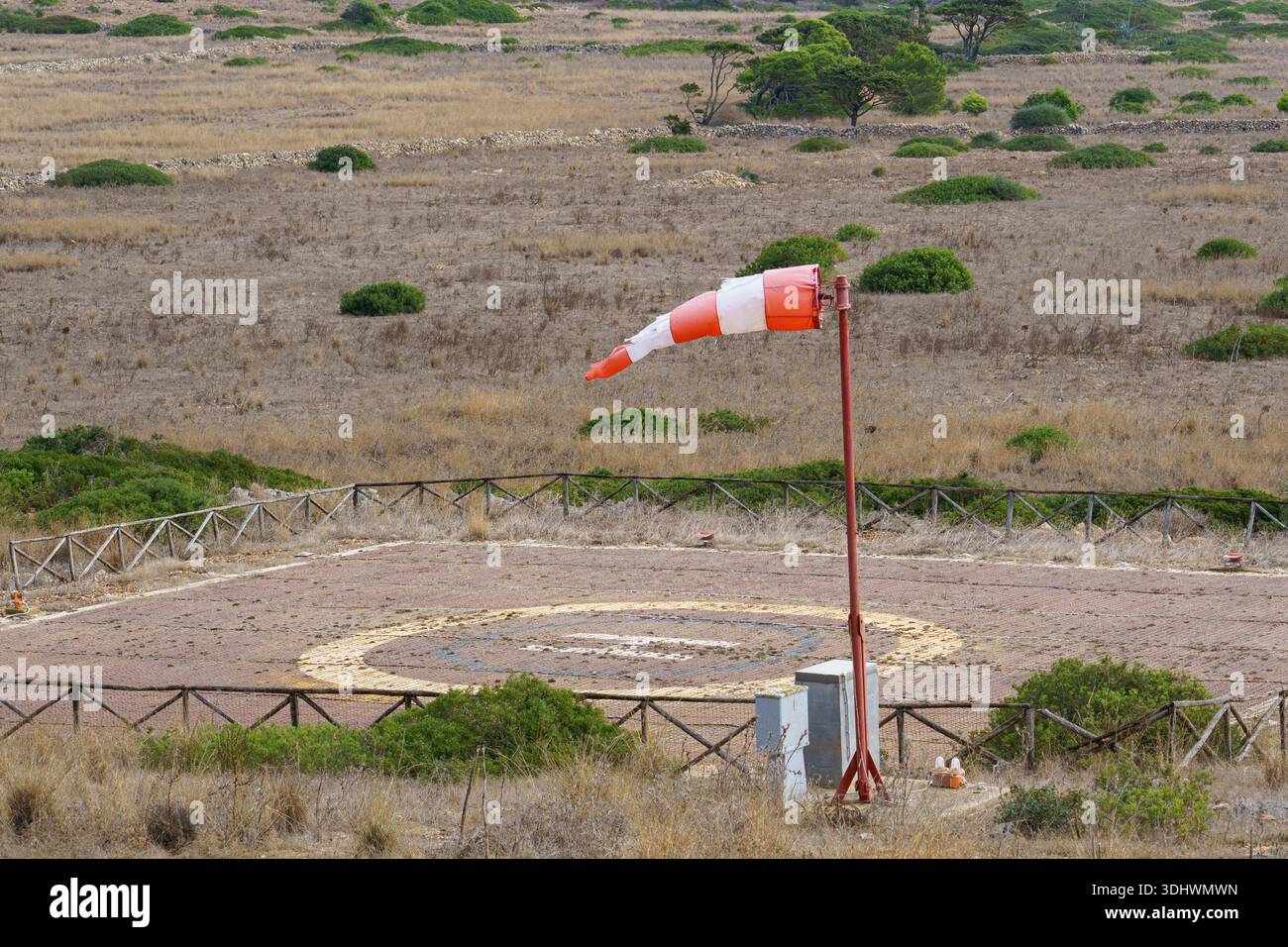 Eine orangefarbene und weiße Windsocke steht neben einem eingezäunten, eigens dafür vorgesehenen Hubschrauberlandeplatz auf einem flachen, trockenen Feld und symbolisiert den Fernzugang, Essential u Stockfoto
