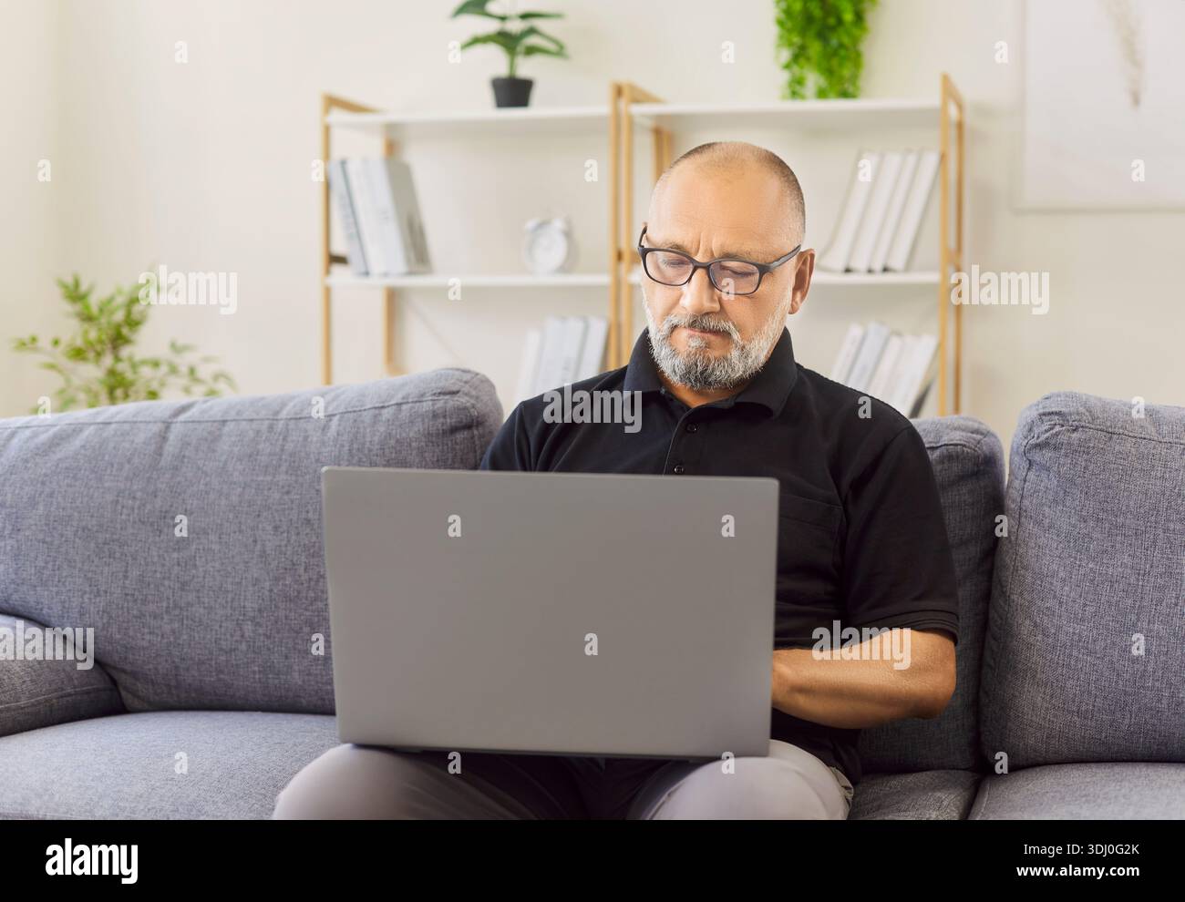 Seriöser älterer Mann, der eine Brille trägt, mit dem Laptop auf einem bequemen Sofa zu Hause Stockfoto