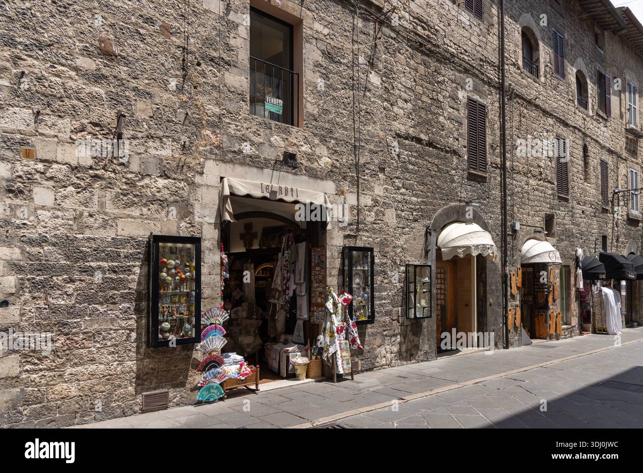 Assisi, Italien - 12. Juni 2025: Typische enge mittelalterliche Assisi-Straße mit Steinfassaden, kleinen Souvenirläden, Blumentöpfen und spiritueller Stille. Stockfoto