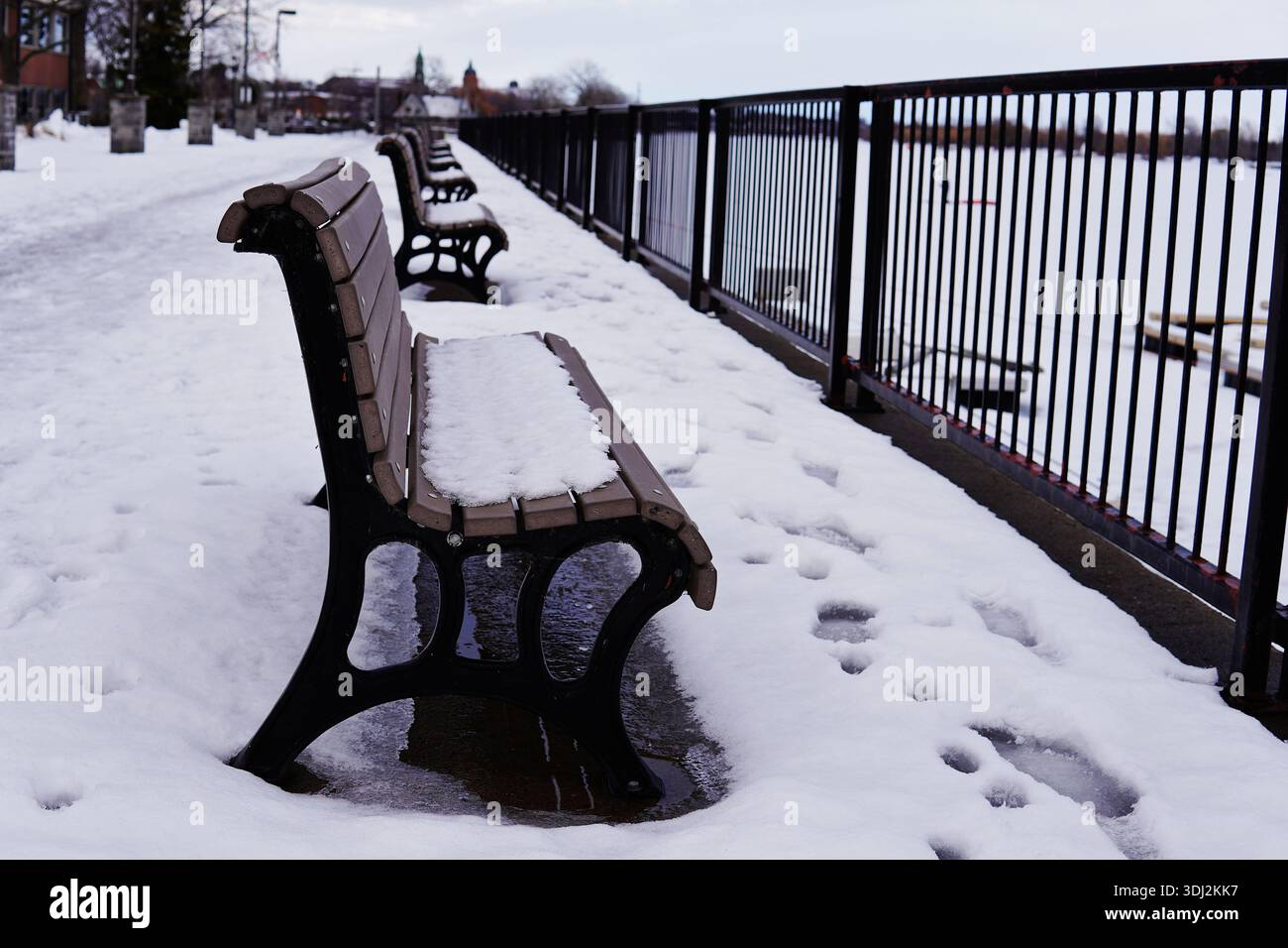 Reihe von schneebedeckten Parkbänken entlang einer Uferpromenade im Winter. Lineare Perspektive mit schwarzem Metallgeländer unter bewölktem Himmel. Stockfoto