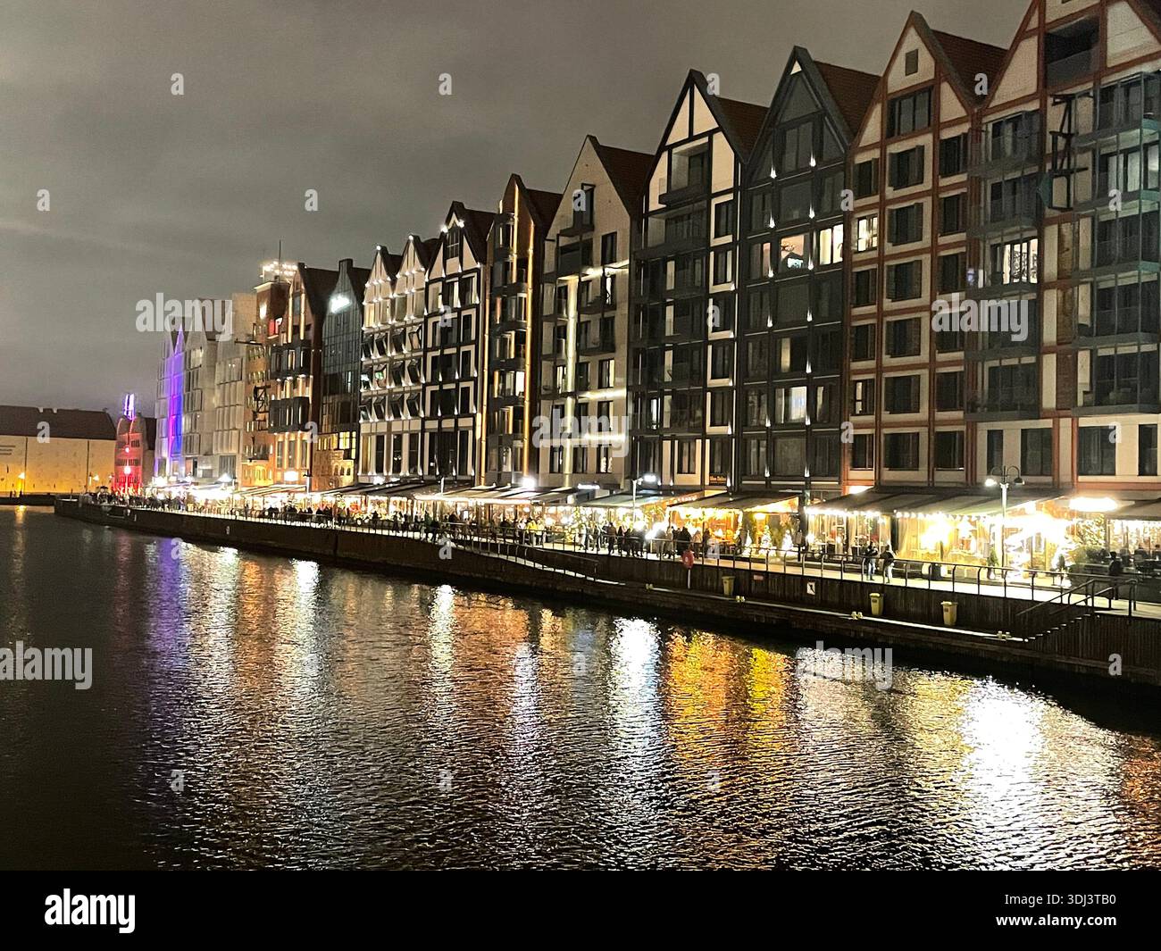 Beleuchtete historische Gebäude am Ufer des Flusses Motława in Gdańsk, Polen, die sich während einer ruhigen abendlichen Stadtszene im Wasser spiegeln - Smartphone-aufgenommenes Stockfoto