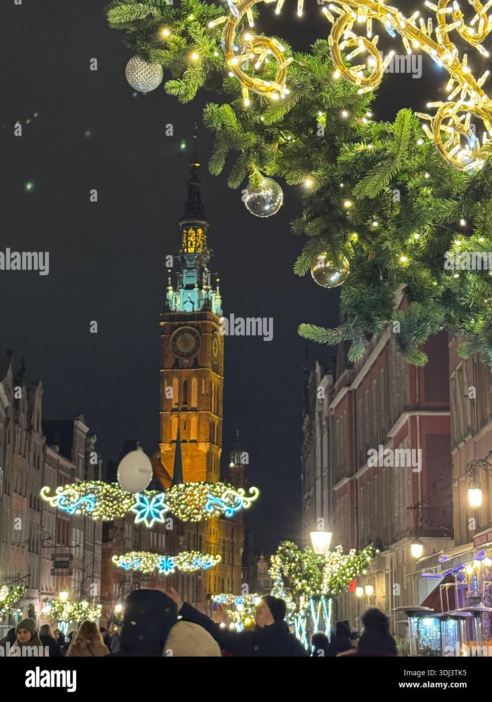 Weihnachtslichter schmücken eine historische Straße mit dem beleuchteten Rathausturm in der Altstadt von Gdańsk bei Nacht. - Smartphone-aufgenommenes Stockfoto