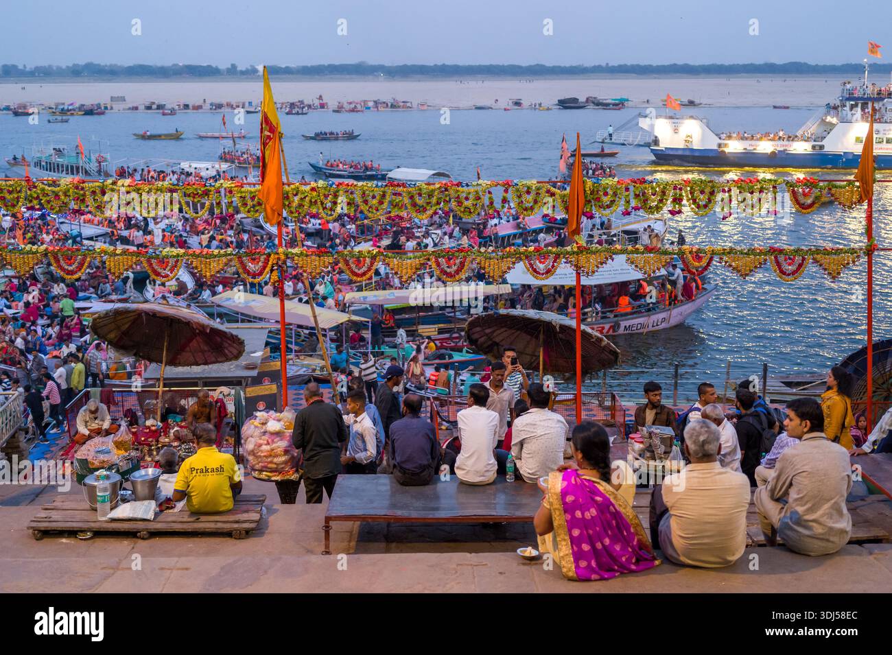 Varanasi, Indien - 17. März 2024: Große Gruppen von Menschen sitzen und stehen auf den Stufen und Plattformen von Dashashwamedh Ghat, mit dekorierten Booten, orangefarbenen Flaggen, Girlanden und Regenschirmen, die während einer Abendveranstaltung entlang des Ganges sichtbar sind. Stockfoto