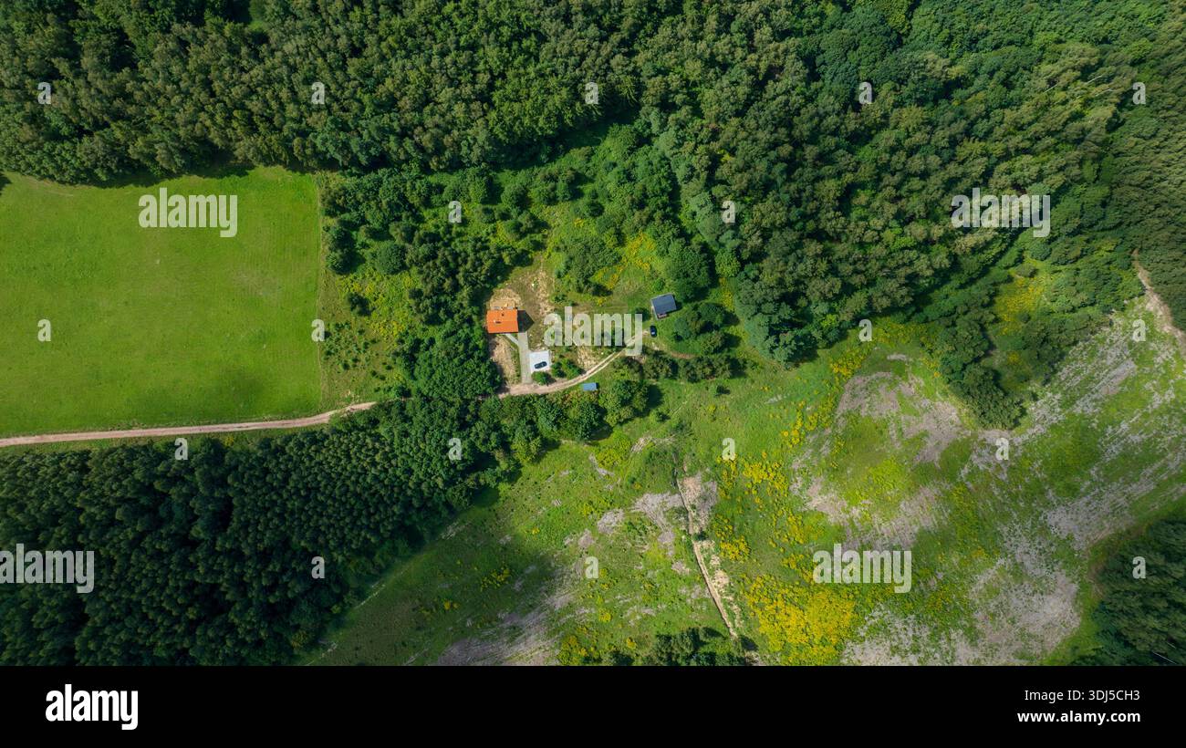 Die Luftaufnahme zeigt ein rotes Dach mit dichtem Wald, eine schmale Schotterstraße, ein Nebengebäude und ein geparktes Auto. Mittagslicht hebt Wiesen, Lichtungen, Stockfoto