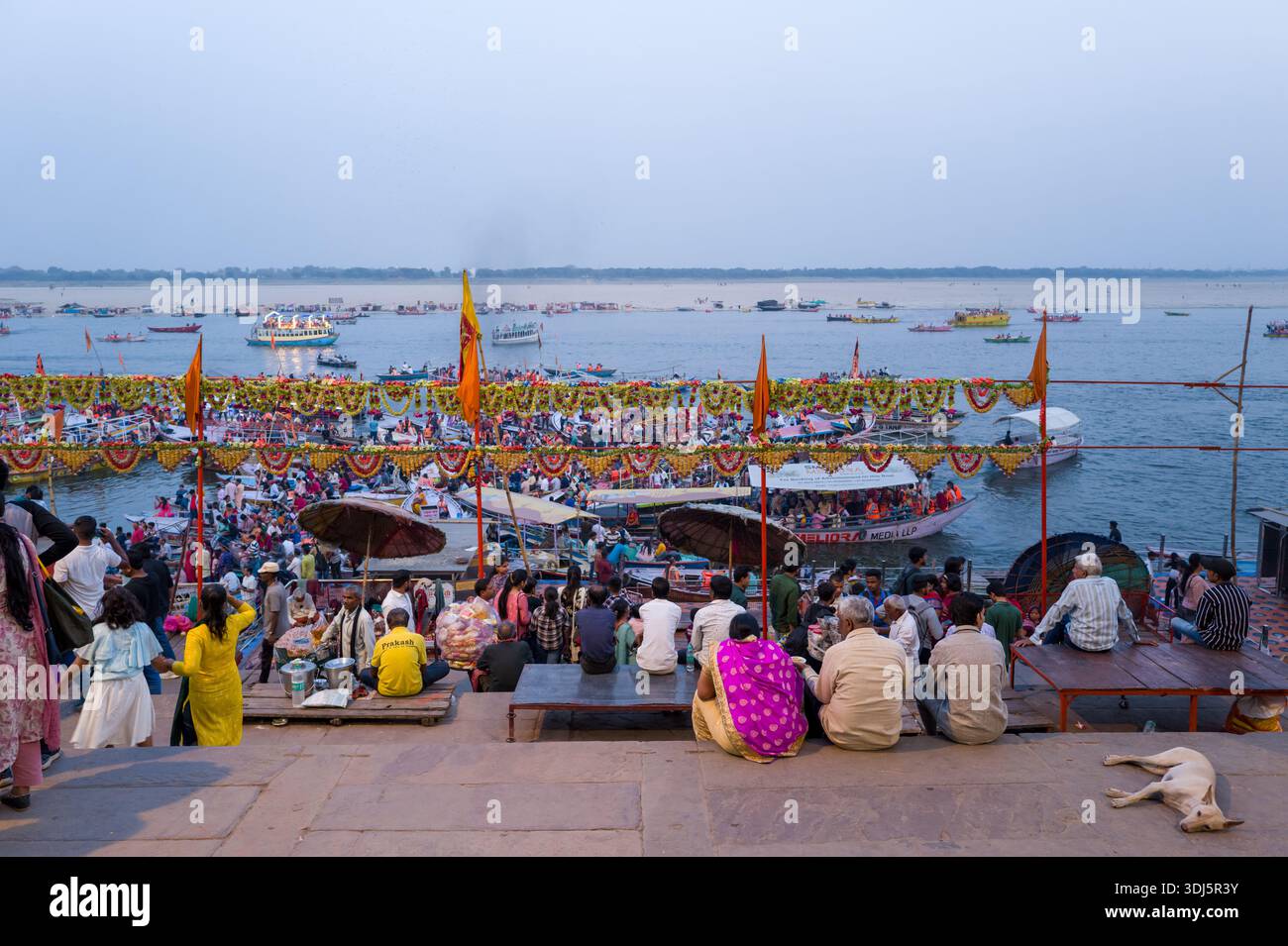 Varanasi, Indien - 17. März 2024: Große Gruppen von Menschen sitzen und stehen auf den Stufen und Plattformen von Dashashwamedh Ghat, mit dekorierten Booten, orangefarbenen Flaggen, Girlanden und Regenschirmen, die während einer Abendveranstaltung entlang des Ganges sichtbar sind. Stockfoto