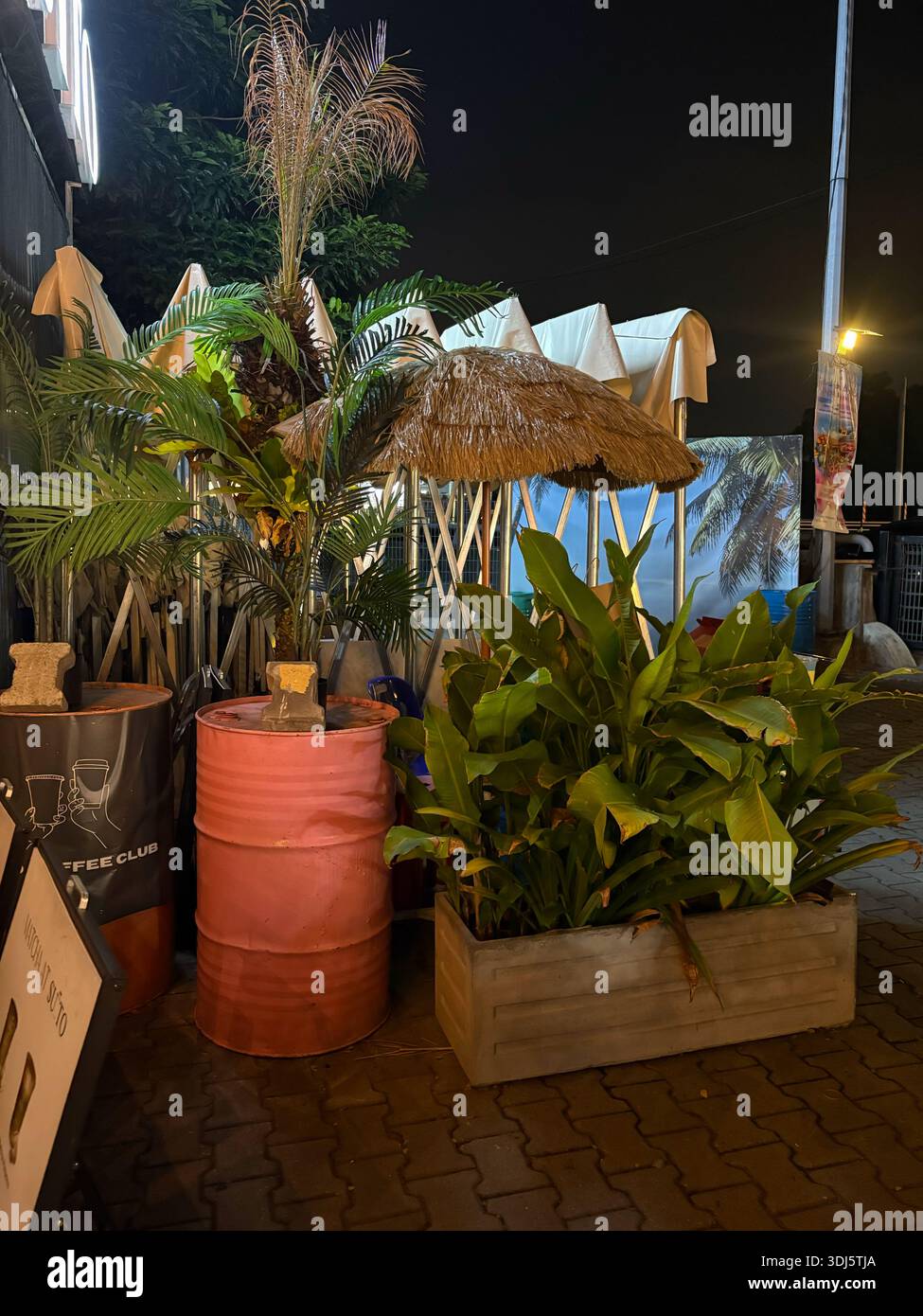 Perak, Malaysia - 24. Januar 2026. Ein abendlicher Sitzbereich im Freien mit tropischen Pflanzen, einem Strohschirm und industriellen Ölfässern als Tische - Smartphone-aufgenommenes Stockfoto