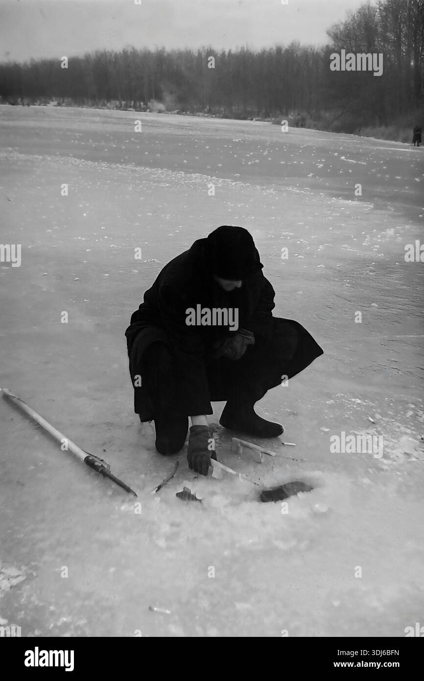 Slowiansk, Region Donezk, Ukraine, UdSSR. 1970er Jahre Ein einsamer Eisfischer hockt in der Nähe eines Lochs auf der gefrorenen Oberfläche eines Flusses. Er trägt einen dunklen Wintermantel und Hut. Ein kleiner Fisch, den er gerade gefangen hat, liegt auf dem Eis in der Nähe seiner Handschuhe. Links vom Fischer liegt ein traditioneller Eismeißel („Peshnya“) auf dem Schnee. Im Hintergrund erstreckt sich eine dunkle Waldlinie entlang des Ufers unter grauem Himmel. Das Bild zeigt die Geduld und die harten Bedingungen des Winterfischens im friedlichen Donbass. Ein archiviertes Schwarzweißfoto. Stockfoto